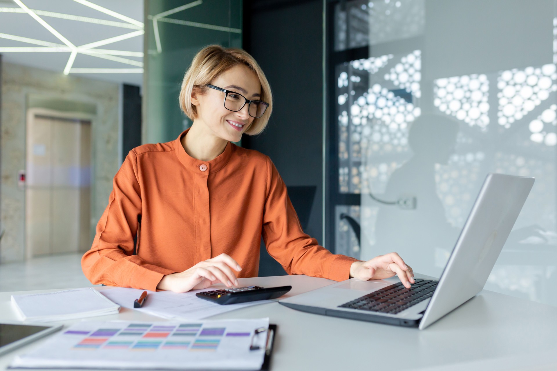 Young female financier with calculator working inside office at workplace, businesswoman behind paper work satisfied smiling, good achievement results, working with contract, accounts and charts